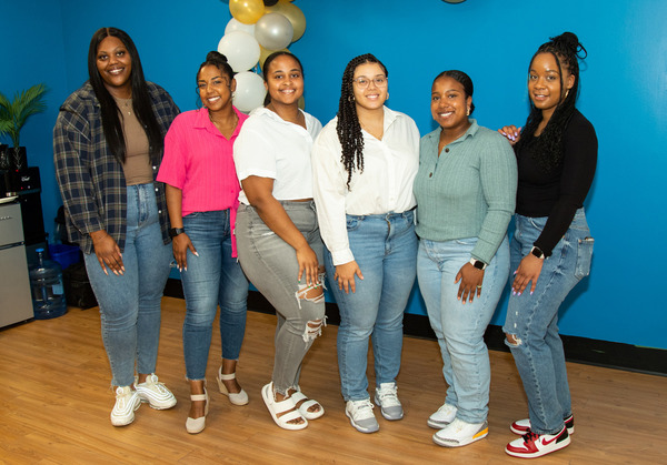 A group of six Black women stand together in front of a blue wall, all smiling broadly.