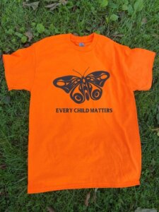 A bright orange t-shirt lies on the grass. The shirt features a black butterfly motif with the words "EVERY CHILD MATTERS" in black beneath.