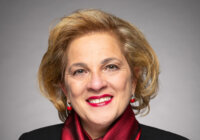 A portrait of a light-skinned woman with short sandy blond hair; she smiles at the camera. She is wearing a red scarf tied at the neck, and matching red lipstick and earrings. Her blazer is black with a silver lapel pin.