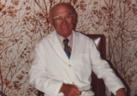 Pearlman in a white doctor's coat seated at a table holding a corded telephone.