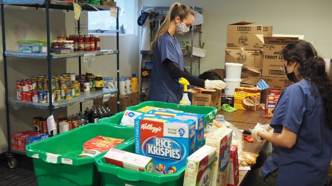 Two people sorting and organizing food at a table with green bins and a shelf behind them.