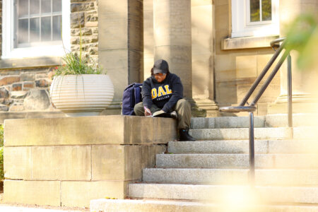 A university student in a DAL hoodie sits on the steps of a stone building on a university campus, writing in a notebook.