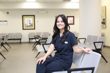 A woman wearing navy blue scrubs sits on a chair in a waiting room
